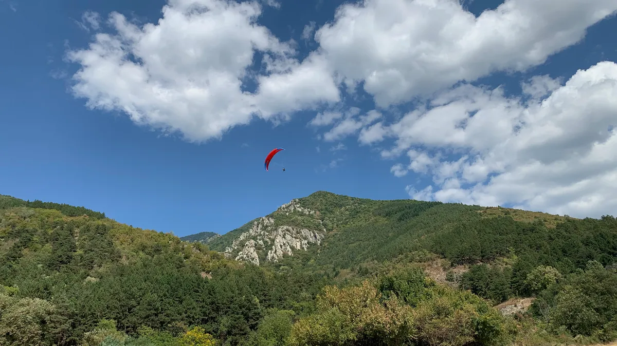 Un parapentiste avec une voile rouge planant haut au-dessus des montagnes boisées et rocheuses de Sopot, en Bulgarie, sous un ciel bleu parsemé de nuages blancs.